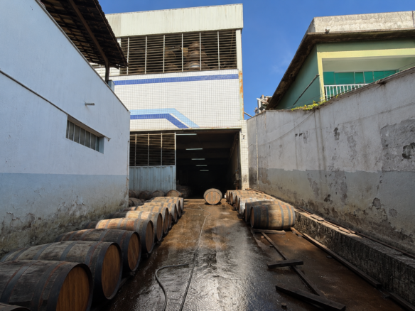 Corredor externo do galpão no Industrial 1, em Belo Horizonte, com fileiras de barris de carvalho e acesso coberto ao fundo